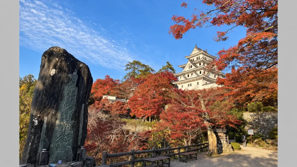 Gujō Hachiman Castle rises above red autumn foliage beside a stone monument in Gifu Prefecture, Japan.